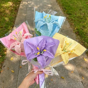 Peut inclure: Quatre bouquets de lys crochetés en rose, bleu, violet et jaune. Chaque bouquet est enveloppé dans du papier coloré et du cellophane, noué avec un ruban portant l'inscription "BEST WISH".