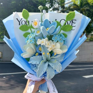 Op de afbeelding: Een handgemaakt gehaakt boeket met blauwe, witte en gele bloemen met groene bladeren. Het boeket is verpakt in blauw papier en vastgebonden met een wit lint. De tekst "You LOVE" is zichtbaar.