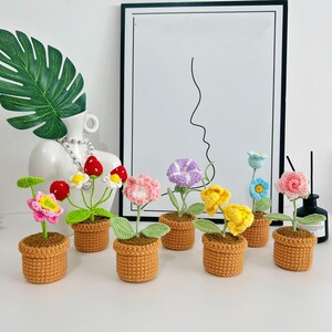 May include: A selection of handmade crochet flowers in small brown pots. The flowers are in various colours, including pink, yellow, purple, and blue, with green leaves. The pots are arranged on a white surface, with a minimalist art print in the background.