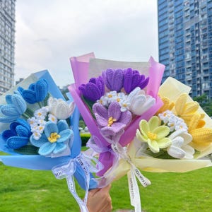 May include: Three bouquets of crocheted flowers in blue, purple, and yellow, wrapped in colorful paper and tied with ribbons. The bouquets feature tulips, daisies, and other flower varieties. The ribbons say "FOR YOU".