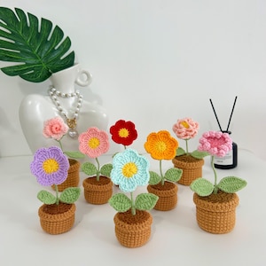 May include: A collection of handmade crochet flowers in small brown pots. Each flower has a different color, including pink, red, orange, blue, and purple, with yellow centers and green leaves. The pots are a warm brown color.