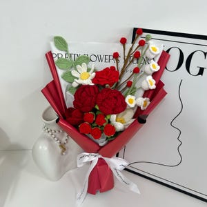 May include: A handcrafted bouquet of crocheted flowers, featuring vibrant red roses, white daisies, and delicate white blossoms. The bouquet is wrapped in red paper and tied with a white ribbon. The text "PRETTY FLOWERS STUDIO" is visible on the paper.