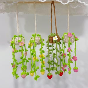 Pode incluir: Jardineiras suspensas de crochê feitas à mão com trepadeiras, flores e frutas. As jardineiras são em tons de bege, marrom, verde, rosa, amarelo e vermelho. As jardineiras suspensas são suspensas em uma haste de madeira.