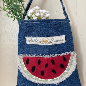 May include: A denim tote bag with a watermelon slice applique and the text "Hello Summer." The bag is filled with white flowers and greenery. The watermelon slice is red with black seeds and a green rind.