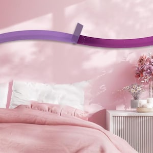 Op de afbeelding: Een moderne, gebogen wandplank in tinten paars en magenta, gemonteerd op een roze muur. De plank bevindt zich boven een bed met witte kussens en een roze dekbed. Een bijzettafel met bloemen en kaarsen is ook zichtbaar.