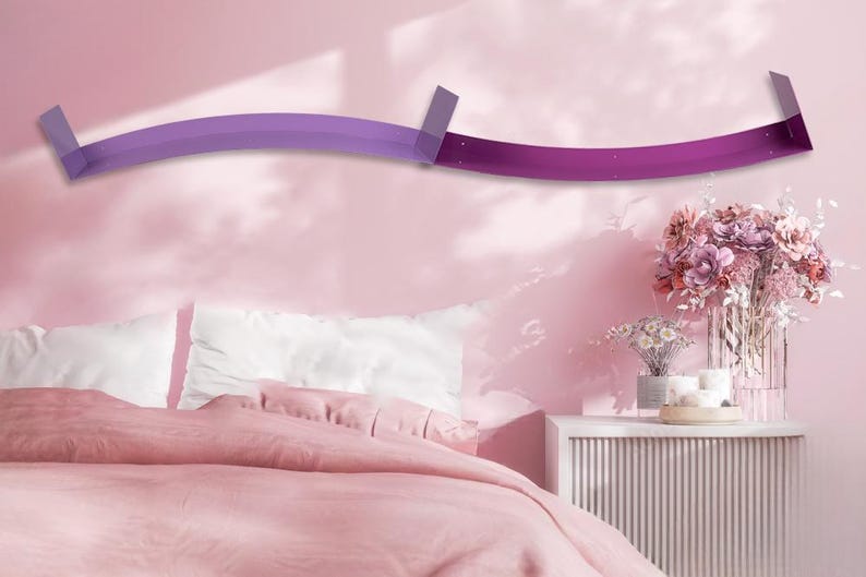 Op de afbeelding: Een moderne, gebogen wandplank in tinten paars en magenta, gemonteerd op een roze muur. De plank bevindt zich boven een bed met witte kussens en een roze dekbed. Een bijzettafel met bloemen en kaarsen is ook zichtbaar.