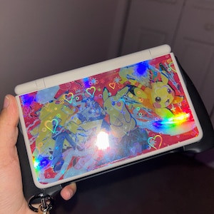 May include: A handheld gaming console with a decorative cover featuring Pikachu and other characters. The cover has a holographic effect with hearts and colorful accents. The console is held in a hand.