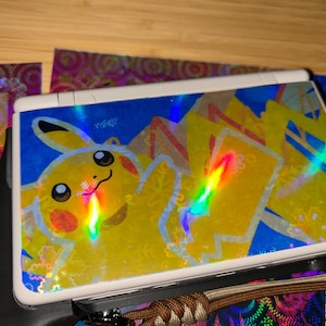May include: A white Nintendo 3DS console with a holographic Pikachu skin. The skin features a yellow Pikachu character on a blue background with lightning bolts. The console is resting on a black surface with colorful accents.