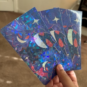 May include: Three holographic stickers with a dark blue background and a repeating design of a crescent moon, stars, and a red and white creature. The stickers have a shiny, iridescent finish.