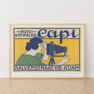 Op de afbeelding: Een ingelijste vintage posterprint met een vrouw die een grootformaatcamera gebruikt. De poster heeft een gele achtergrond met blauwe en zwarte tekst, waaronder "FOTO-ARTIKELEN" en "Capi". De vrouw draagt een groene top en heeft donkerblauw haar.