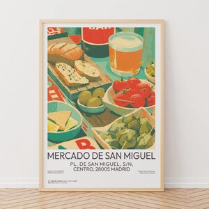 May include: A framed art print featuring a vibrant illustration of a Spanish market scene. The artwork showcases a table laden with food, including bread, tomatoes, olives, and artichokes, alongside a glass of beer. The text "MERCADO DE SAN MIGUEL" is displayed.