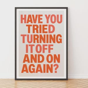 Op de afbeelding: Een ingelijste wandkunstprint met de tekst HAVE YOU TRIED TURNING IT OFF AND ON AGAIN? in dikke, oranje, schreefloze hoofdletters, gecentreerd op een lichtgrijze achtergrond. De lijst is dun, donker en rechthoekig van vorm.