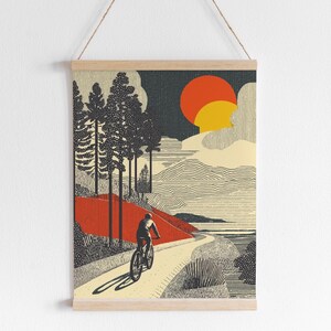 May include: A framed art print depicting a cyclist on a winding road. The artwork features a stylized landscape with trees, a red hill, and a setting sun in shades of orange and yellow. The print is mounted on a wooden frame and suspended by a string.