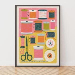 Op de afbeelding: Een ingelijste kunstdruk met een kleurrijke illustratie van diverse klossen naaigaren in tinten roze, oranje, geel en blauwgroen, samen met een donkergroene schaar, alles tegen een gespleten roze en gele achtergrond.