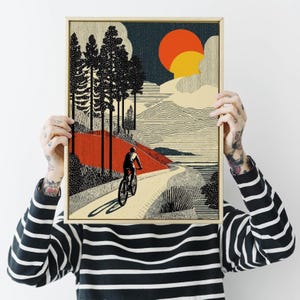 May include: A framed art print depicting a cyclist on a winding path through a landscape. The artwork features a red and beige color palette with black trees, a setting sun, and a textured style. The print is held up by a person.