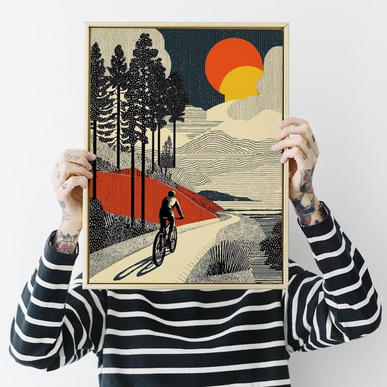 May include: A framed art print depicting a cyclist on a winding path through a landscape. The artwork features a red and beige color palette with black trees, a setting sun, and a textured style. The print is held up by a person.