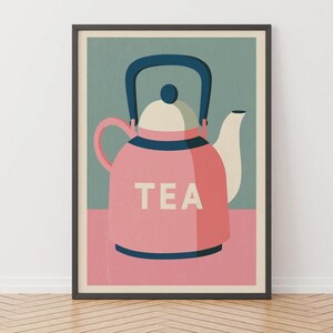 Op de afbeelding: Een ingelijste kunstdruk met een gestileerde illustratie van een roze theepot met een donkerblauw handvat en dekselknop. Het woord TEA staat in witte hoofdletters op de voorzijde van de theepot, geplaatst tegen een gedempte groene achtergrond.