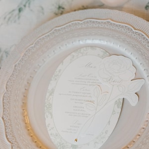 Op de afbeelding: Een ovaalvormig papieren menu met een lichtgroene bloemenrand en een uitgestanste roosillustratie aan de rechterkant. Het menu bevat bovenaan het woord Menu, gevolgd door de gangen, en ligt op een wit decoratief bord.