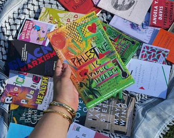 Könnte beinhalten: Eine Sammlung von Büchern mit verschiedenen Titeln, darunter "Palestine is Public Health" und "Black Marxism." Die Bücher sind auf einem schwarz-weiß gemusterten Tuch angeordnet, mit einer Hand, die ein Buch hält. Die Buchumschläge sind farbenfroh.