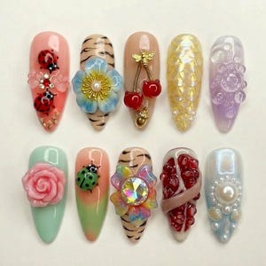 May include: A collection of ten artificial nails with various designs. The nails feature embellishments such as ladybugs, flowers, cherries, and pearls. Colors include pink, green, yellow, and purple. Some nails have a tiger stripe pattern.