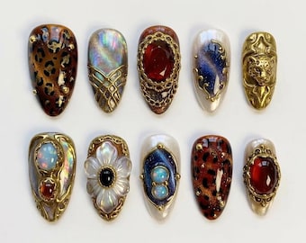 Könnte beinhalten: Eine Sammlung von zehn mandelförmigen Press-on-Nägeln mit aufwendigen Designs. Die Nägel weisen Leopardenmuster, schillernde Effekte, Edelsteine und Goldfiligran auf. Jeder Nagel hat einzigartige Verzierungen mit einer Vielzahl von Texturen und Farben, darunter Rot, Blau und Gold.
