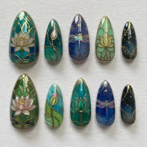 May include: A collection of ten false nails, each with an almond shape and detailed artwork. The designs include lotus flowers, dragonflies, and starry night scenes, all framed with gold accents. The colour scheme features teal, blue, green, and gold.