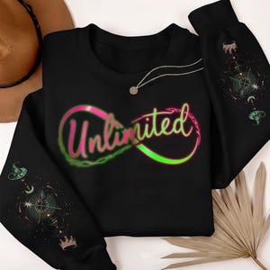 Può includere: Felpa nera con la scritta "Unlimited" in un design a simbolo infinito rosa e verde. Le maniche presentano grafiche celesti e stregonesche. Una collana d'argento con un piccolo ciondolo è drappeggiata sulla felpa.