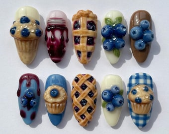 May include: A collection of ten artificial nails with various blueberry-themed designs. The nails feature miniature blueberry muffins, pie, and other dessert-inspired art. Colors include blue, brown, white, and shades of purple.