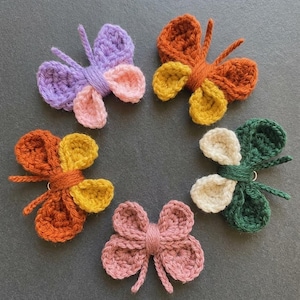Può includere: Una collezione di sei ornamenti a farfalla all'uncinetto in vari colori, tra cui viola, arancione, giallo, verde e rosa. Ogni farfalla ha ali e antenne dettagliate, disposte in un motivo circolare su una superficie grigia.