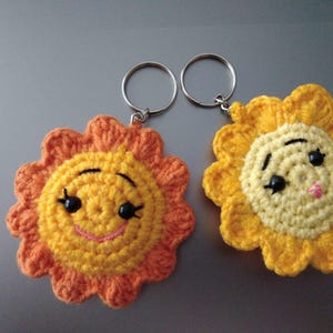 May include: Two handmade crochet sun keychains. One sun is orange and yellow, the other is yellow. Both have smiling faces with black eyes and a pink mouth. The keychains are attached to silver key rings.