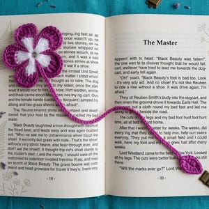 May include: A crocheted bookmark rests in an open book. The bookmark features a purple and white flower design with a long, matching purple stem. A small metal charm is attached to the end of the stem.