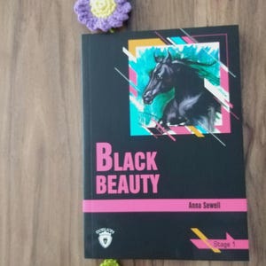 May include: A hardcover book titled "Black Beauty" by Anna Sewell, featuring a black horse illustration on the cover. The book is black with pink and yellow accents. A purple and yellow crochet flower and a green crochet leaf are next to the book.