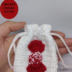 Op de afbeelding: Een kleine, witte gehaakte zak met trekkoord en een rood-wit bloemmotief. De tas heeft een gestructureerd steekpatroon en een decoratieve rood-witte bloemenapplicatie. De tekst "CROCHET PATTERN ENG PDF" staat in een rode cirkel.