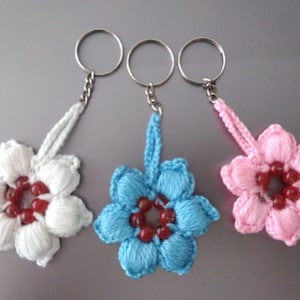 May include: Three handmade flower keychains in white, blue, and pink. Each flower is crocheted with a central cluster of red beads. The keychains have silver-colored metal rings and chains for attaching to keys or bags.