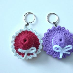 May include: Two handmade crochet keychains. One is red with a white scalloped edge and a white bow. The other is purple with a white bow. Both have silver key rings attached. The keychains are round and have a spiral design.
