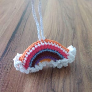 Puede incluir: Adorno de arcoíris de ganchillo hecho a mano con un lazo blanco para colgar. El arcoíris presenta capas de naranja, rosa, rojo, azul, morado y amarillo. El borde inferior está adornado con volantes blancos.