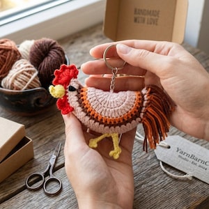 Può includere: Un portachiavi a forma di gallo all'uncinetto nei toni del rosa, arancione e marrone. Il gallo ha una cresta rossa e zampe gialle. Il portachiavi è tenuto in mano, con una ciotola di filato e una piccola scatola nelle vicinanze. L'etichetta recita "YarnBarnCo. Studio Est. 2024."