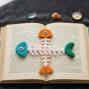 Puede incluir: Un marcapáginas de ganchillo con forma de espina de pescado con detalles en naranja, azul y verde, colocado en un libro abierto. El marcapáginas presenta un diseño de esqueleto de pez blanco con detalles coloridos en forma de ojo. También se ve una brújula y piedras decorativas.