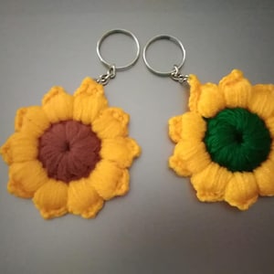 Puede incluir: Dos llaveros de girasol hechos a mano. Cada llavero presenta un girasol tejido con pétalos amarillos y un centro marrón o verde. Los llaveros tienen anillos metálicos plateados para sujetar a llaves o bolsos.