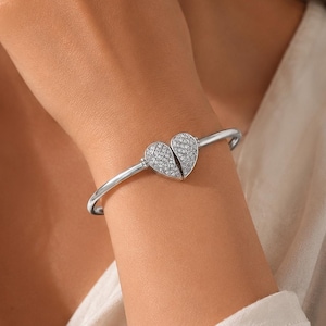 Könnte beinhalten: Ein silberfarbenes Armband mit einem zentralen herzförmigen Anhänger, der aus zwei Hälften besteht. Das Herz ist mit zahlreichen kleinen, klaren, funkelnden Steinen besetzt, die die gesamte Oberfläche des Anhängers bedecken und einen Glanzeffekt erzeugen.