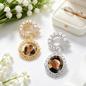 Può includere: Due spille con dettagli di perle e strass e ciondoli con foto, una dorata e una argentata. Sono visibili anche un bouquet di fiori bianchi e un portagioie con due anelli. Le spille presentano un design a ghirlanda con un fiocco.