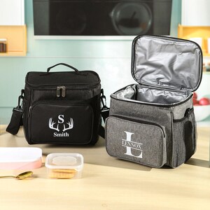 May include: Two insulated lunch bags. One is black with a white deer antler design and the name "Smith". The other is gray with the name "Lennox". Both have a top handle, shoulder strap, and front pocket. The gray bag is open, revealing a silver interior.