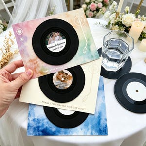 May include: Wedding favors featuring a black vinyl record design. The favors have a photo of the couple in the center and the text "Harry & Rebecca October 16, 2026". The background is a watercolor design with the text "THANKS FOR MAKING THIS DAY A HIT!"
