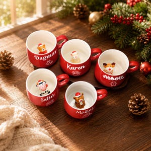 Könnte beinhalten: Fünf rote Keramiktassen mit weißem Innenraum, jede mit einer kleinen 3D-Tierfigur mit Weihnachtsmütze am Boden. Die Tassen sind mit den Namen Dagmar, Karen, Abbott, Eaton und Mabel in weißer Schreibschrift und weihnachtlichen Symbolen personalisiert.