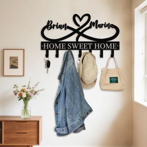 May include: Black metal wall-mounted key and coat rack with the names "Brian" and "Marina" and the phrase "HOME SWEET HOME". A denim jacket, straw hat, and tote bag hang from the hooks. A framed picture and flowers are on a wooden cabinet.