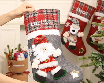 May include: A gray Christmas stocking with a red plaid cuff, personalized with the name "Lucilla". The stocking features a Santa Claus applique and a gold star. Other stockings with names are in the background.