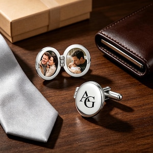 May include: A pair of silver-toned oval cufflinks. One cufflink is open, revealing two circular photo inserts: one showing a couple and the other showing a person holding a baby. The second cufflink is closed, displaying the black engraved letters AG.
