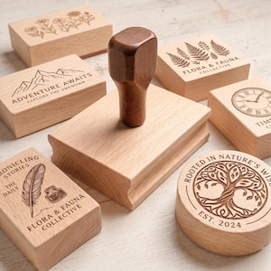 May include: A selection of wooden stamps with engraved designs and text. The stamps display botanical illustrations, mountain scenes, and phrases such as "Adventure Awaits" and "Flora & Fauna Collective." A wooden stamp handle is also visible.