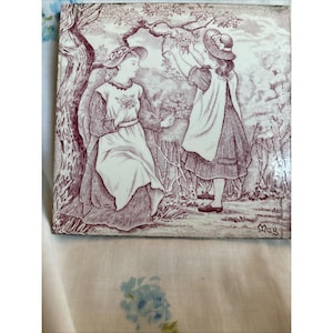 May include: Sepia-toned ceramic tile featuring a garden scene with two figures. One figure is seated, holding flowers, while the other reaches for branches. The tile has a glossy finish and a decorative border. The word "May" is visible in the corner.