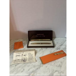 Könnte beinhalten: Ein Vintage-Sheaffer-Stiftset in einer braunen Box, mit einem goldfarbenen Stift und einem passenden Bleistift. Das Set enthält ein orangefarbenes Lederetui, eine kleine orangefarbene Karte und Anweisungen. Der Stift wird auf einer Marmoroberfläche präsentiert.